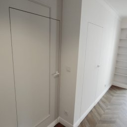 Gładko.pl - Białe, ukryte drzwi wkomponowane w ścianę z dekoracyjnymi listwami przypodłogowymi i parkietem ułożonym we wzór jodełki.