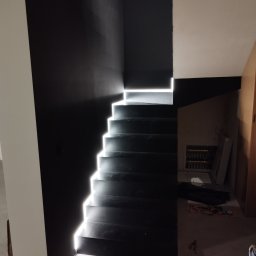 Gładko.pl - Nowoczesne, czarne schody z podświetleniem LED, kontrastujące z białą ścianą, widoczne elementy instalacji elektrycznej w tle.