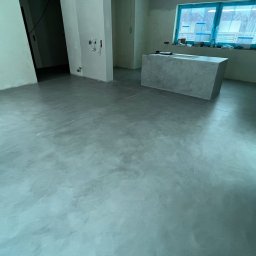 Gładko.pl - Szara, błyszcząca posadzka z mikrocementu w przestronnym wnętrzu, widoczne surowe ściany z instalacjami elektrycznymi i betonowa wyspa kuchenna pod oknem.