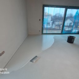 MicroForm Studio - Wylewka samopoziomująca w trakcie realizacji. Widoczna szpachelka i wiadro z masą. Okno zabezpieczone folią. Świeża, jasna wylewka na podłodze w nowym pomieszczeniu.