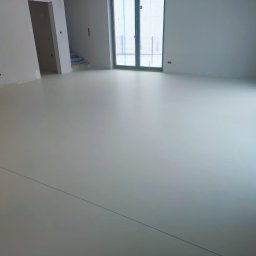 MicroForm Studio - Jasna, gładka posadzka w nowym wnętrzu. Widoczne ściany przygotowane do malowania, okno zabezpieczone folią. Minimalistyczny design, świeżo wylana podłoga.