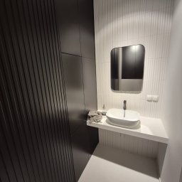 MicroForm Studio - Nowoczesna łazienka z umywalką nablatową, lustrem w zaokrąglonej ramie i ciemną zabudową meblową z ryflowanymi frontami. Białe płytki z delikatnym wzorem.