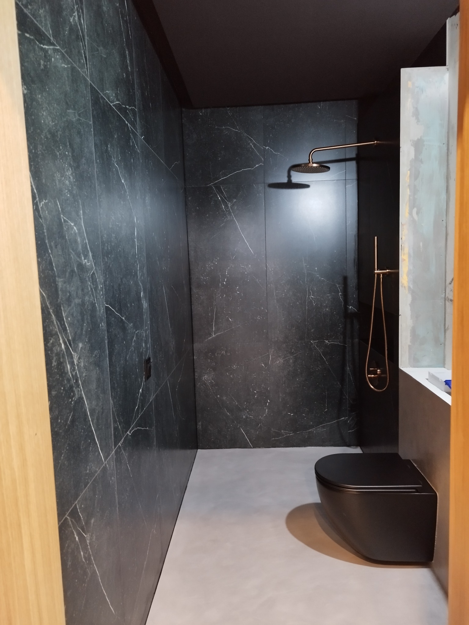 Nowoczesna łazienka z czarnymi płytkami imitującymi marmur, złotą armaturą i podwieszaną toaletą w minimalistycznym stylu, oświetlona punktowym światłem.