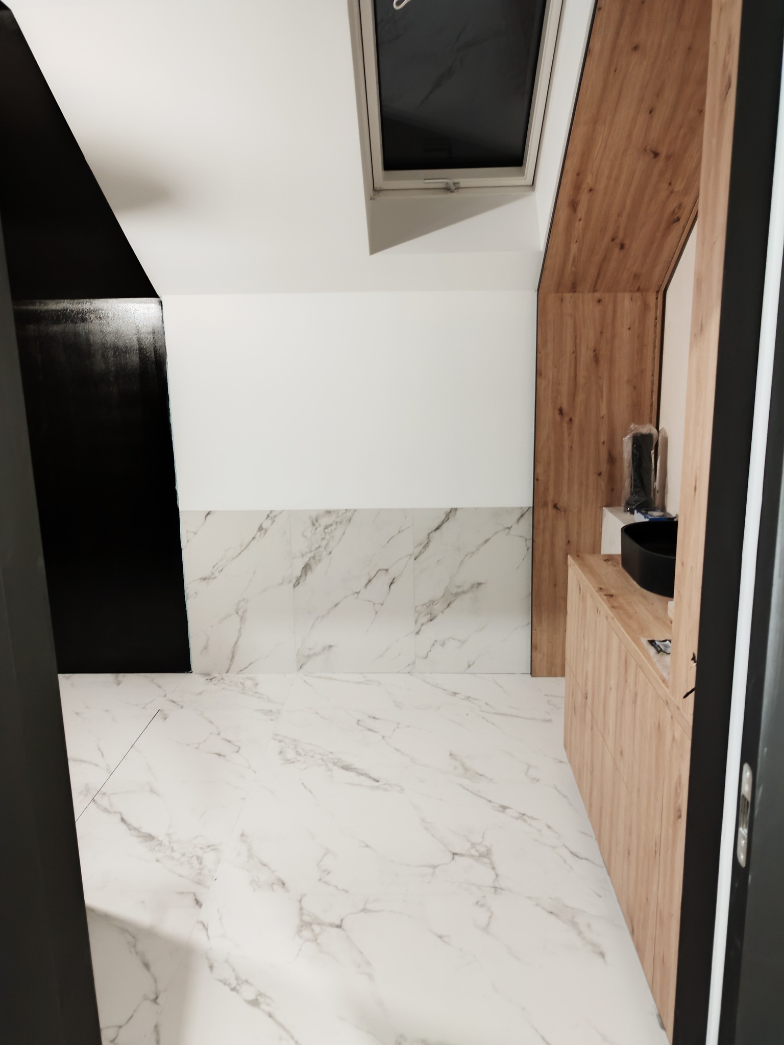 Nowoczesna łazienka z białymi marmurowymi płytkami podłogowymi i ściennymi, czarnymi akcentami na ścianach i drewnianą zabudową umywalki, doświetlona oknem dachowym.