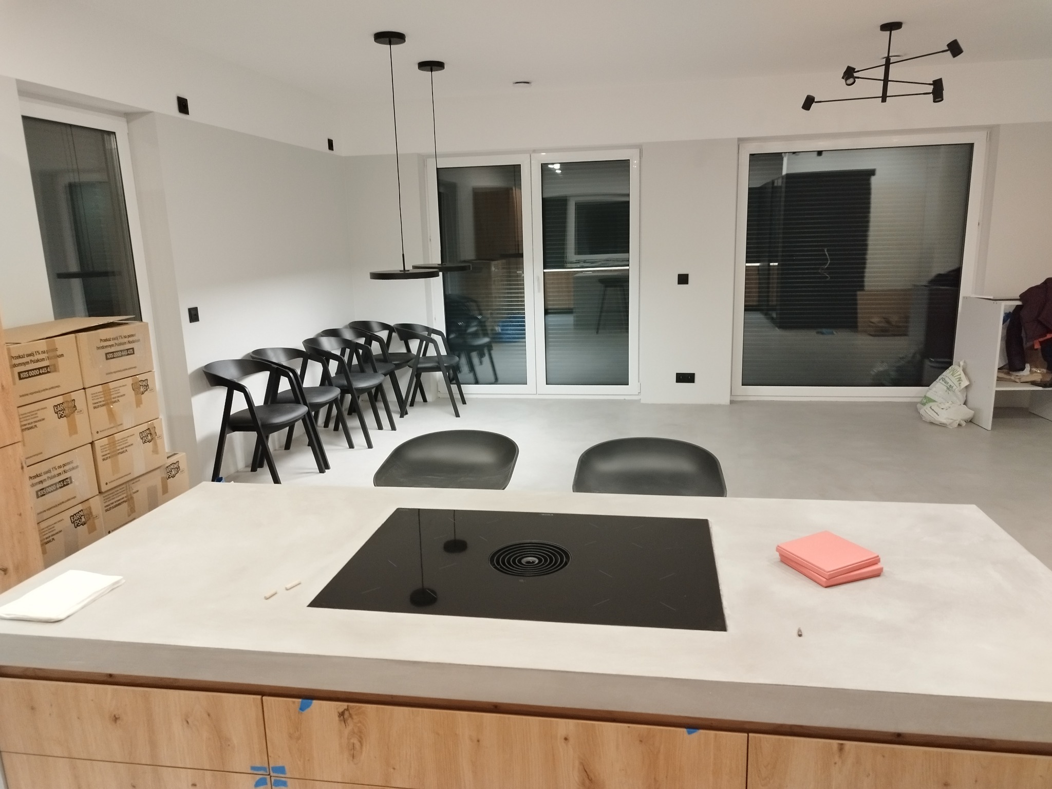 Wnętrze nowoczesnej kuchni w trakcie wykańczania, widoczny blat wyspy z wbudowaną płytą indukcyjną i pochłaniaczem, krzesła w tle, kartony po sprzęcie AGD i surowe ściany.