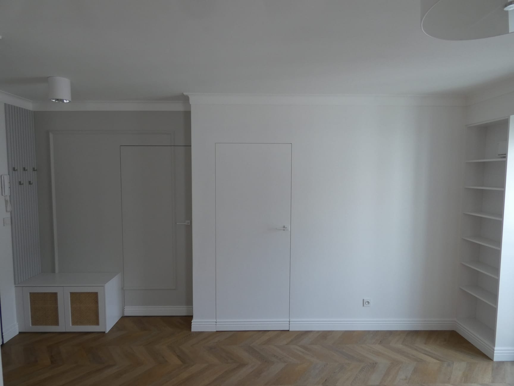 Jasne wnętrze z drewnianą podłogą w jodełkę, białe ściany z listwami przypodłogowymi, zabudowana szafka z rattanowymi frontami, ukryte drzwi i biały regał.