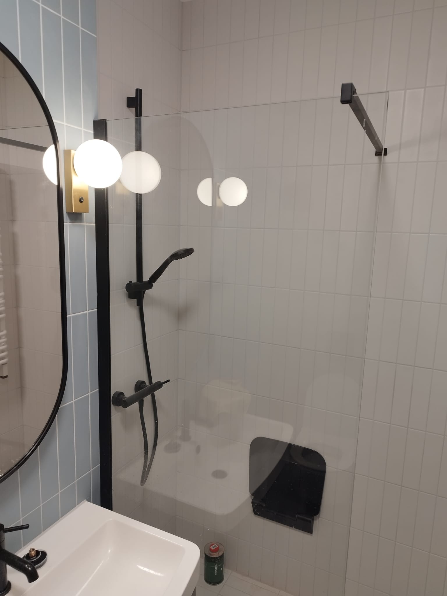 Nowoczesna łazienka z czarną armaturą, szklaną kabiną prysznicową i okrągłym lustrem z oświetleniem kulowym, wykończona płytkami w odcieniach bieli i błękitu.