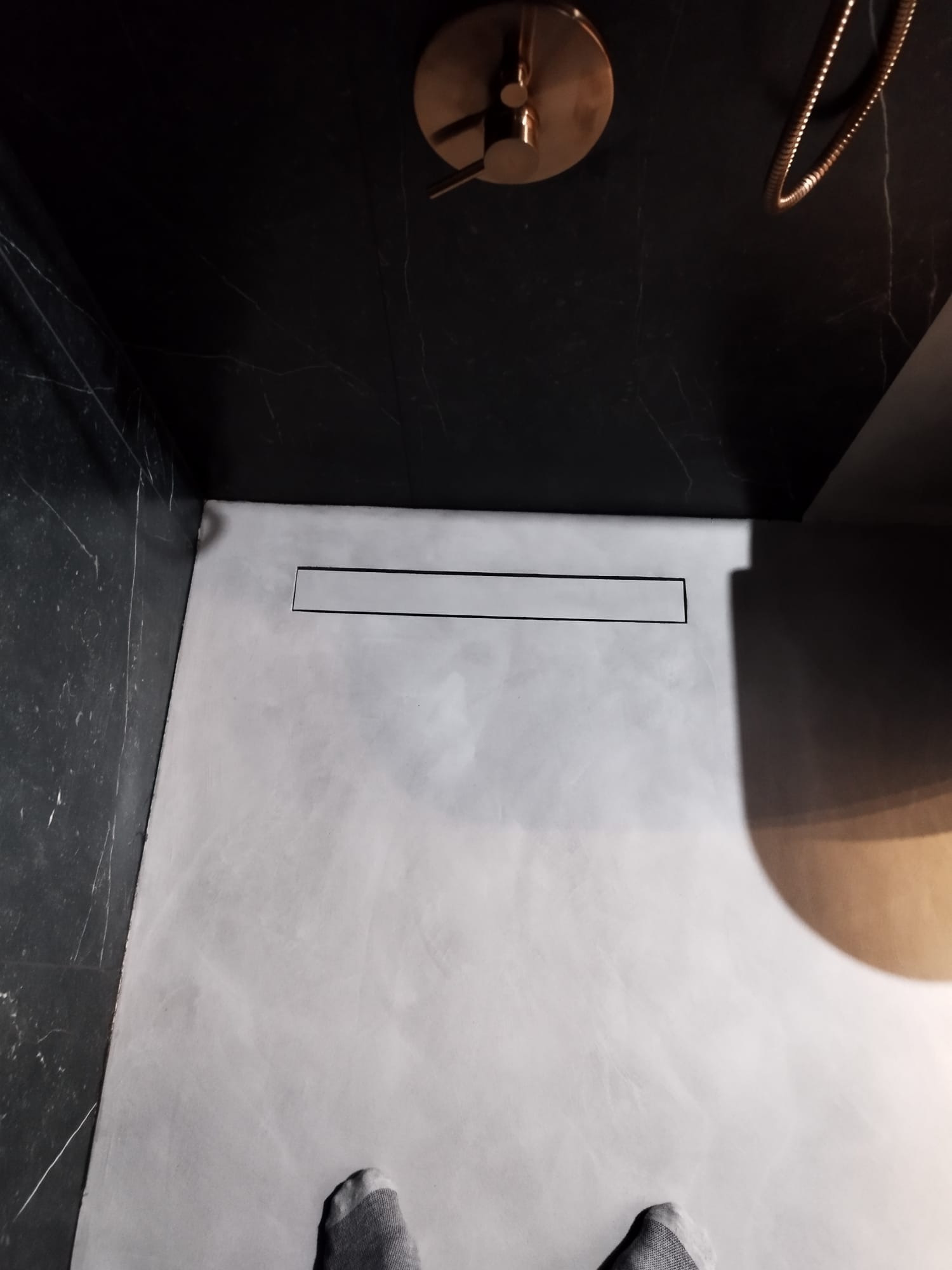 Nowoczesna kabina prysznicowa z szarym wykończeniem podłogi, czarnymi ścianami imitującymi marmur, odpływem liniowym i miedzianą armaturą, widoczne stopy w skarpetkach.