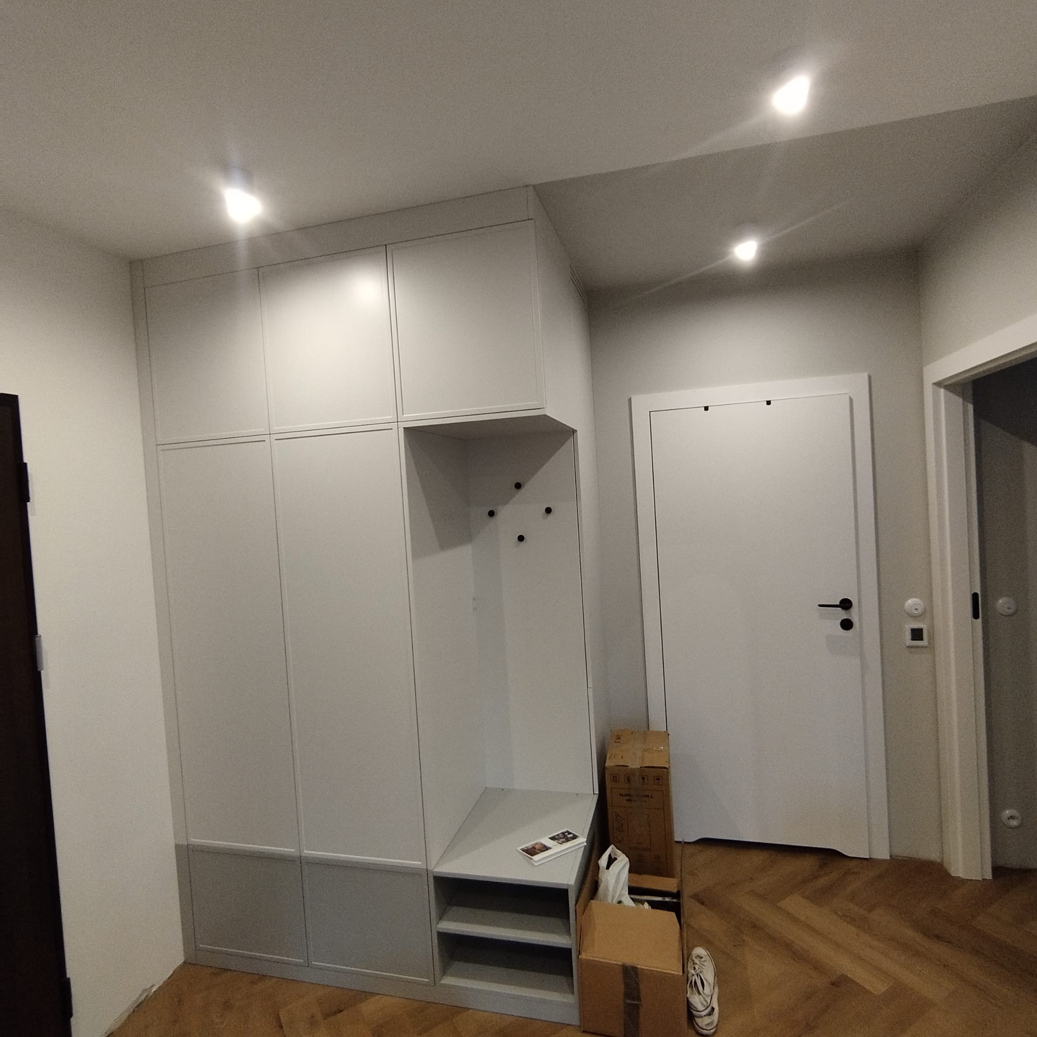 Biała zabudowa meblowa w przedpokoju z siedziskiem i wieszakami, na podłodze parkiet w jodełkę, obok kartony i buty.