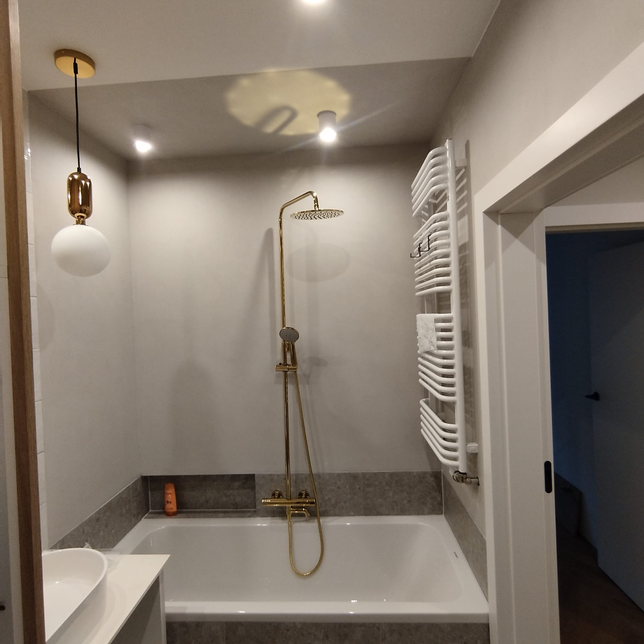 Nowoczesna łazienka z wanną obudowaną szarymi płytkami, złotą armaturą prysznicową i wiszącym grzejnikiem drabinkowym.