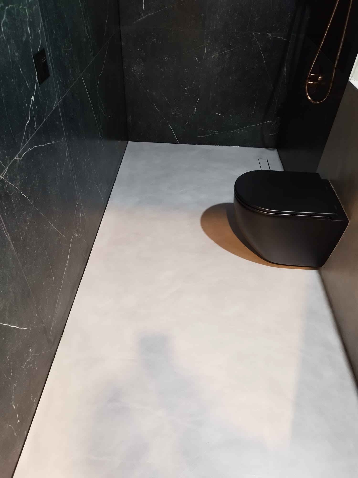 Nowoczesna łazienka z czarną armaturą i szarym mikrocementem na podłodze, ściany wykończone czarnym marmurem z białymi żyłami.