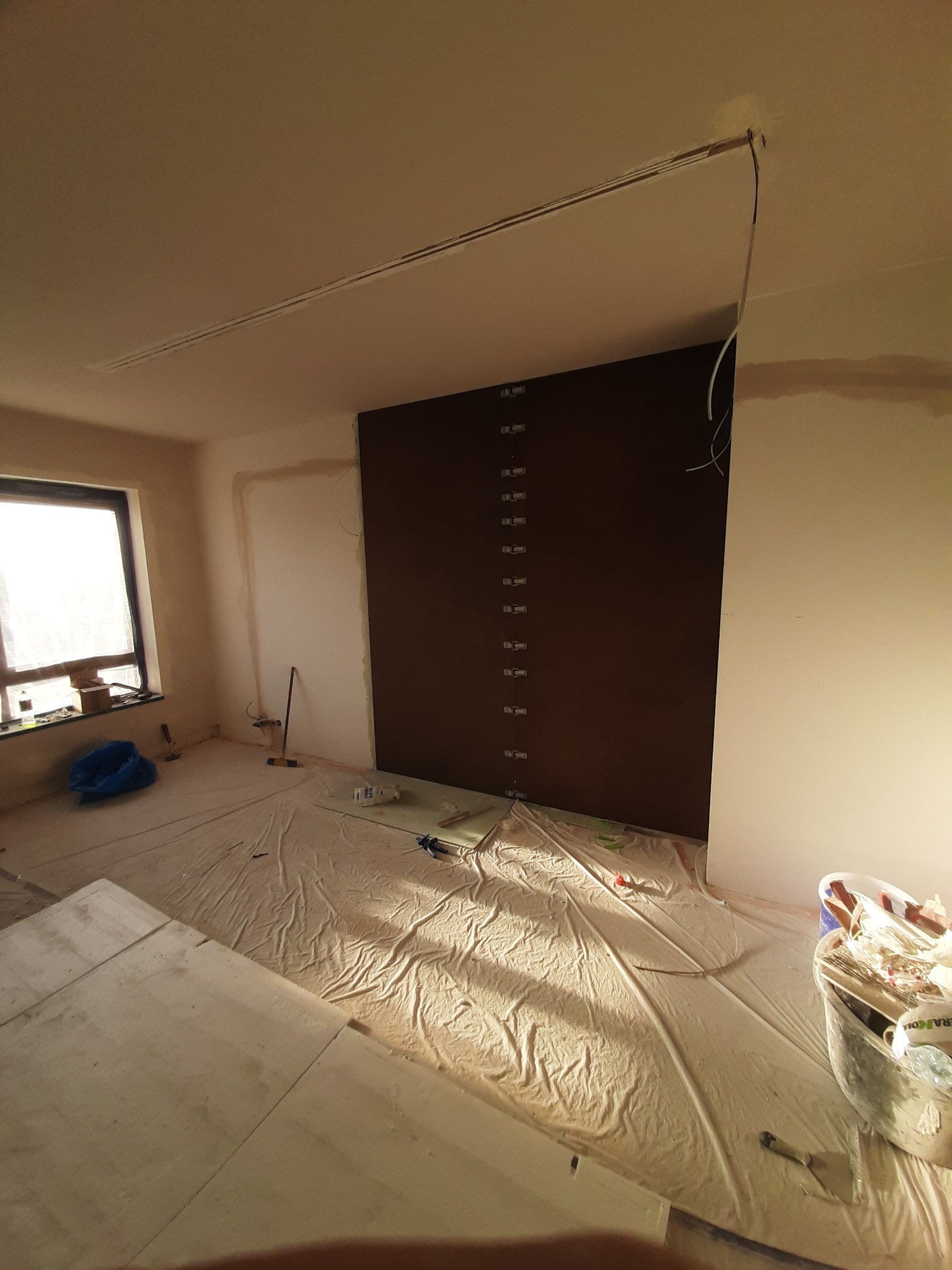 Pokój w trakcie remontu: zabezpieczona folią podłoga, ściany przygotowane do malowania, widoczne ślady po szpachlowaniu, brązowa zabudowa z pionowym rzędem uchwytów po lewej stronie ściany, otwór...