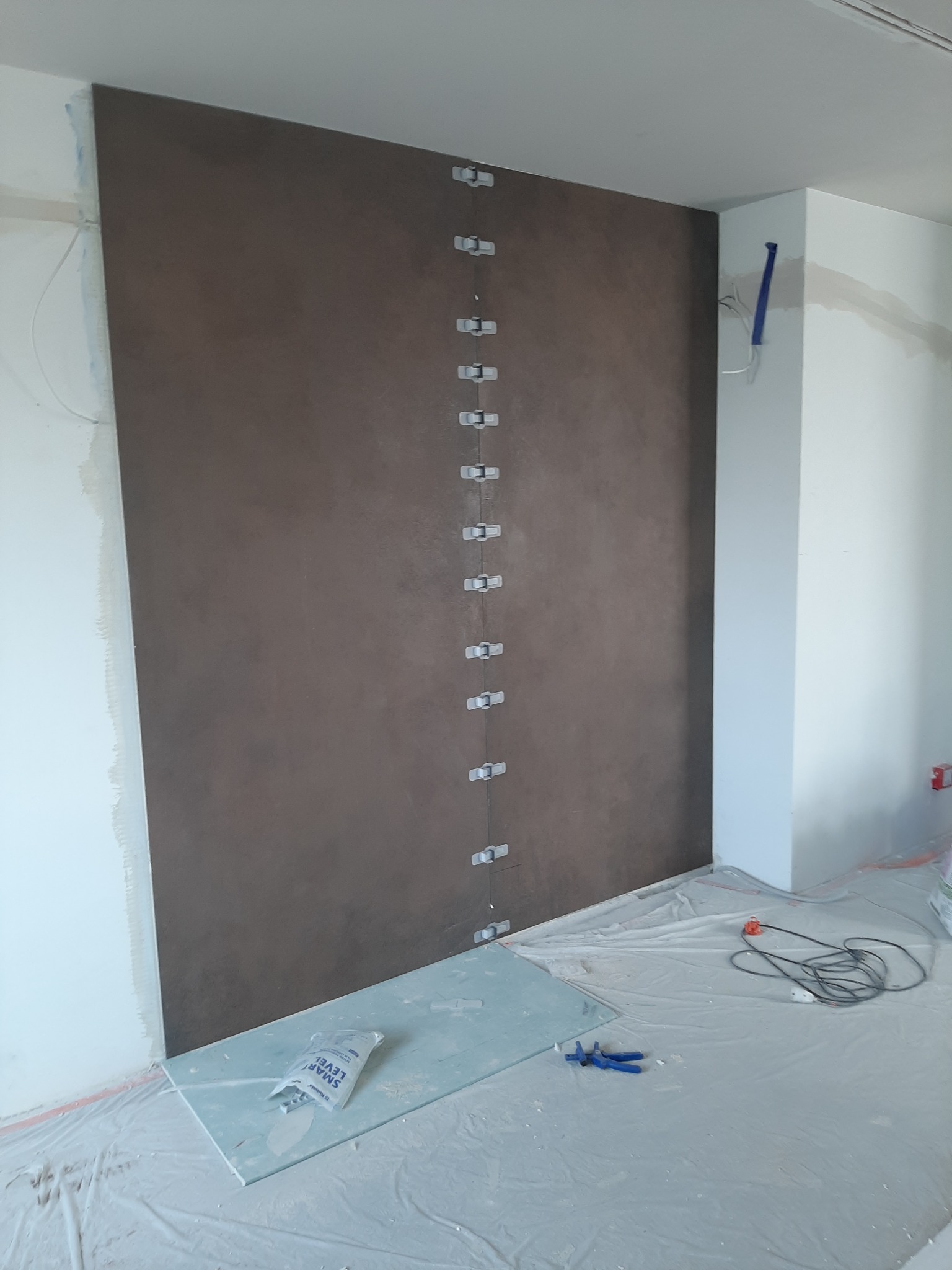 Brązowa ściana z widocznymi łącznikami poziomującymi, zabezpieczona folią podłoga, w trakcie prac wykończeniowych.