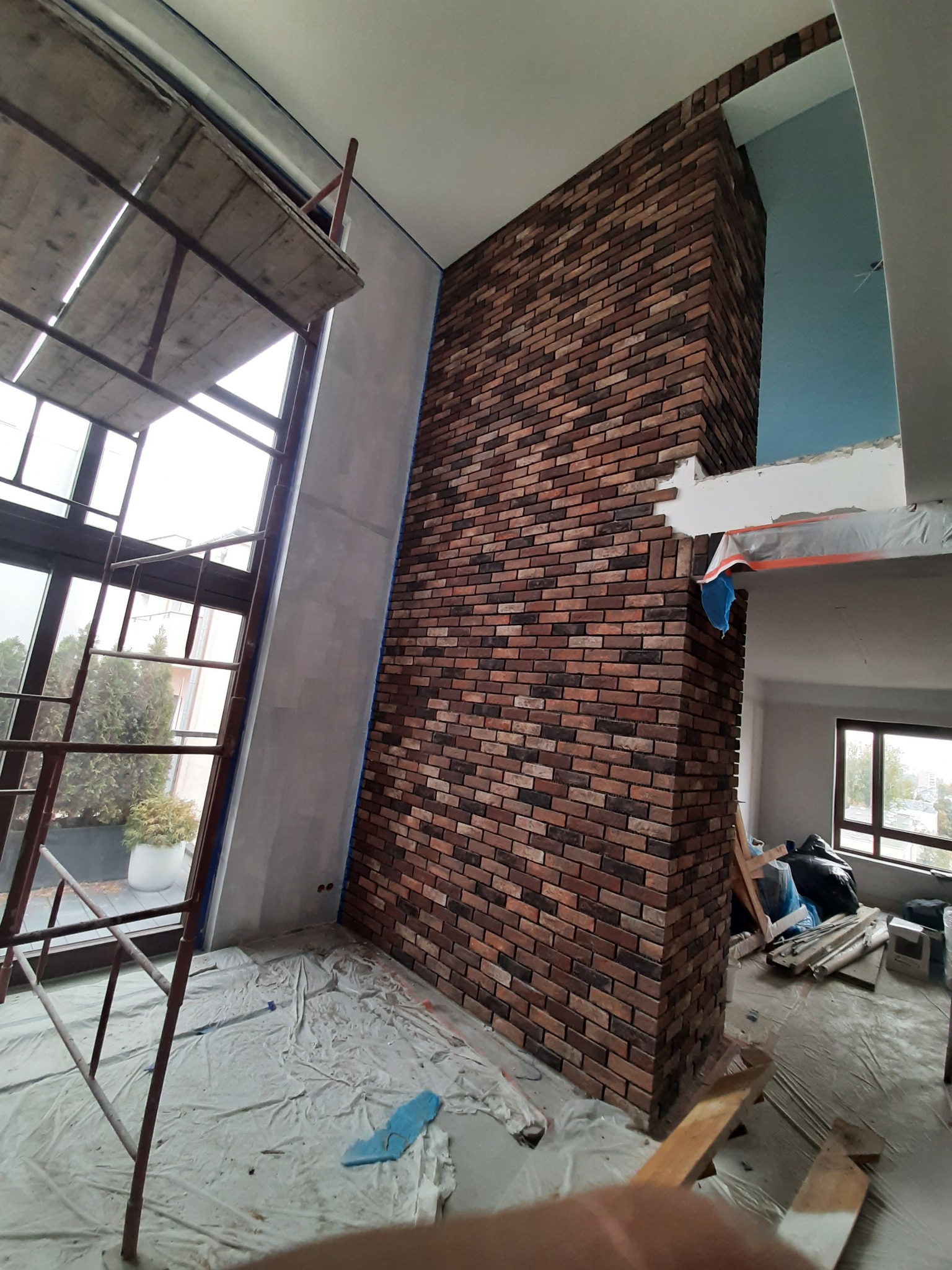 Wnętrze w trakcie prac: częściowo wykończona ściana z cegieł dekoracyjnych, rusztowanie przy oknie, zabezpieczona folią podłoga i widoczne materiały budowlane.