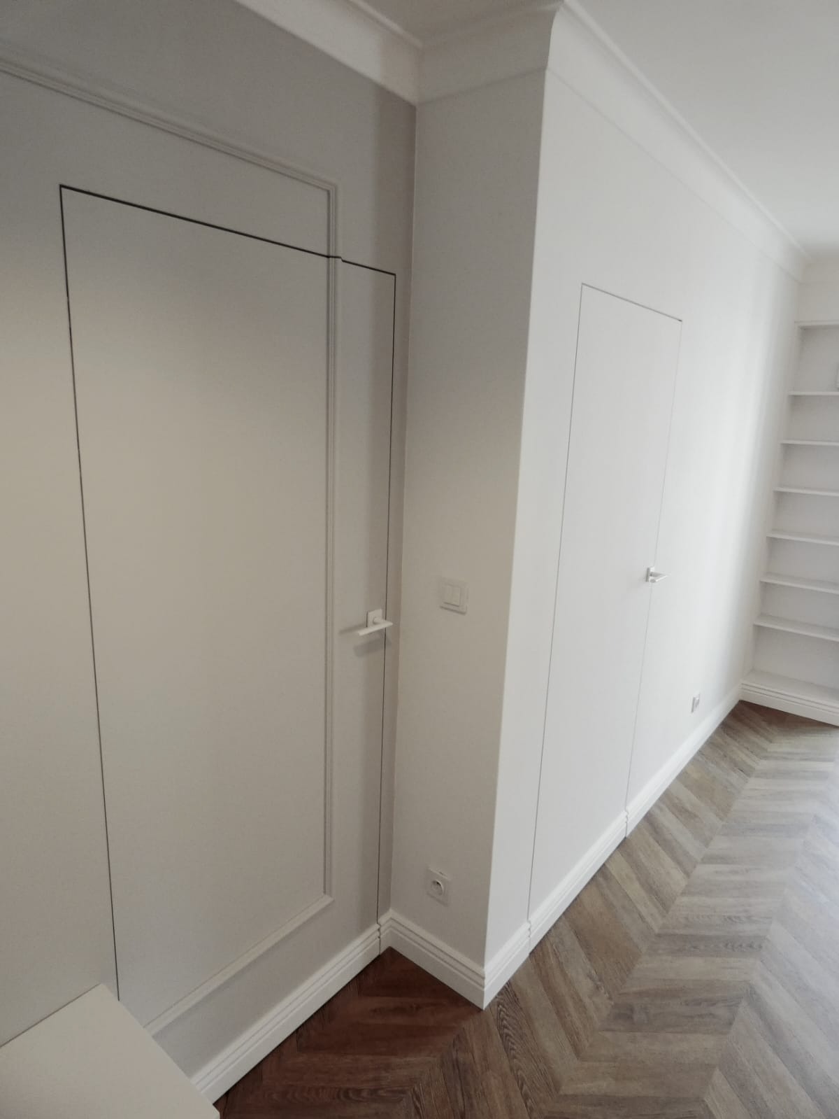 Białe, ukryte drzwi wkomponowane w ścianę z dekoracyjnymi listwami przypodłogowymi i parkietem ułożonym we wzór jodełki.