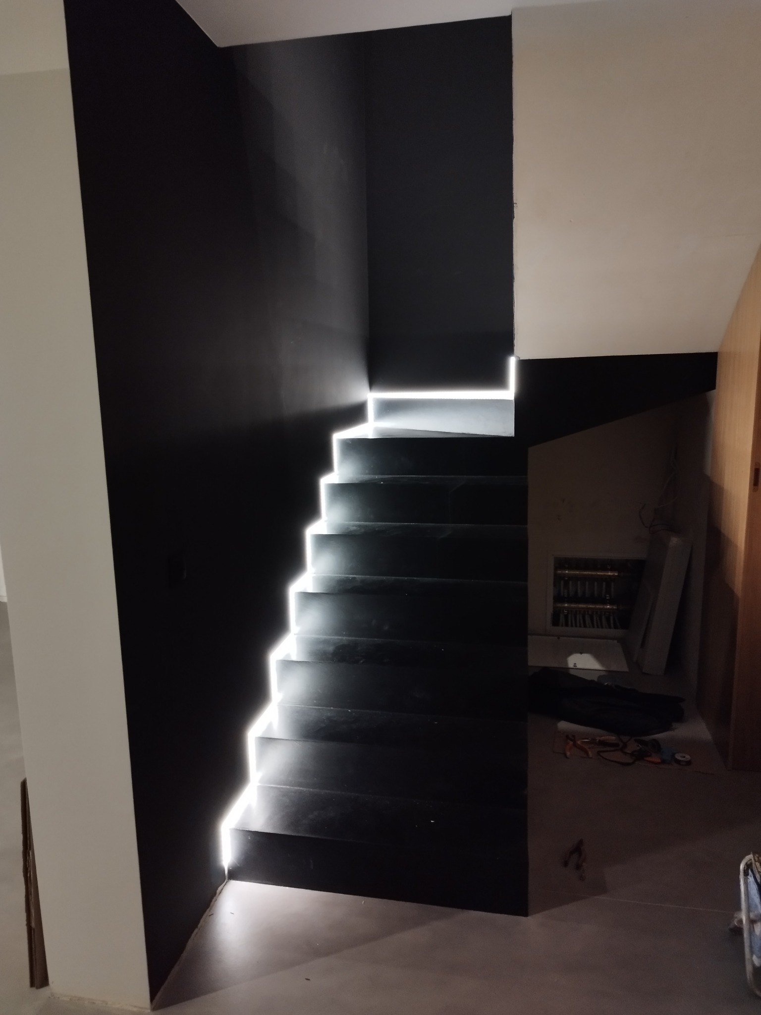 Nowoczesne, czarne schody z podświetleniem LED, kontrastujące z białą ścianą, widoczne elementy instalacji elektrycznej w tle.
