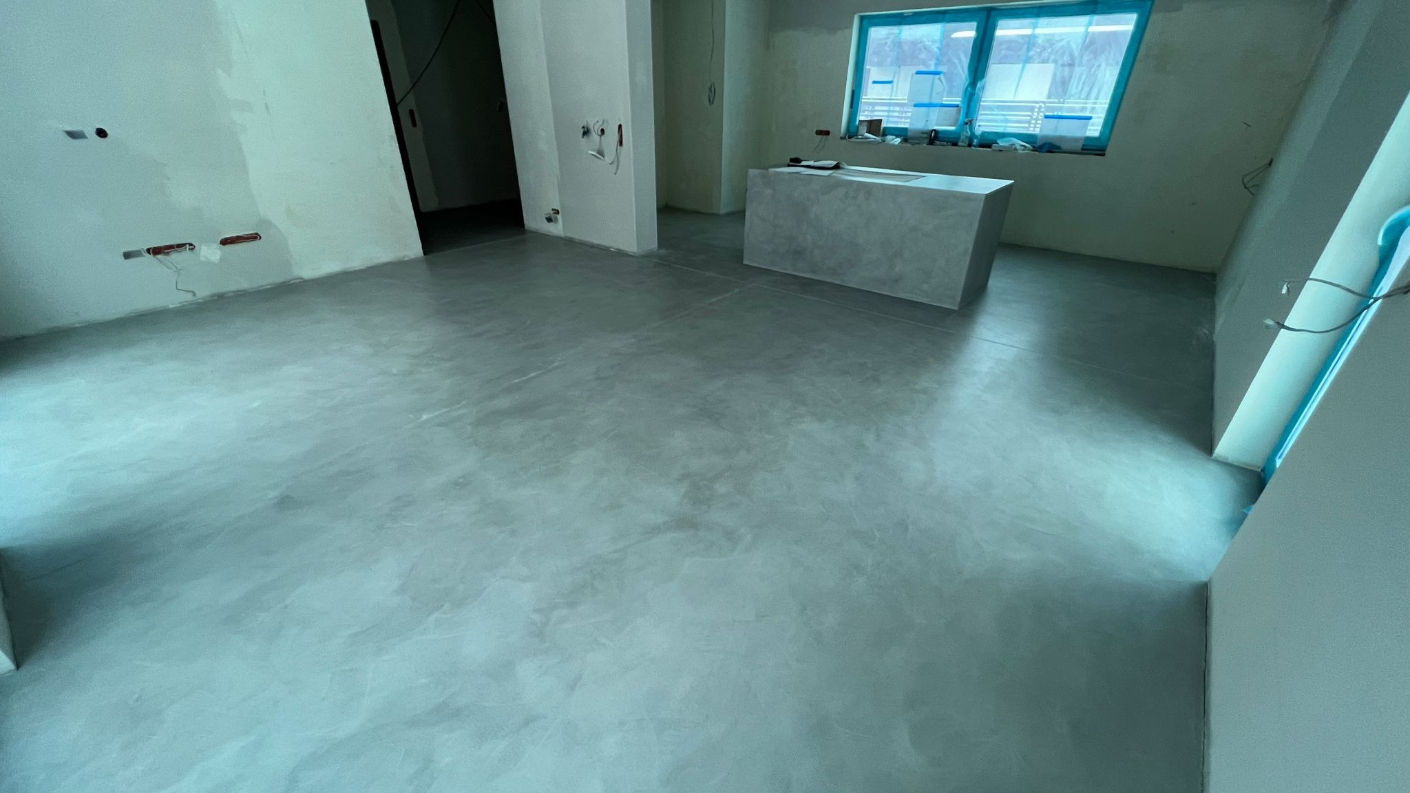 Szara, błyszcząca posadzka z mikrocementu w przestronnym wnętrzu, widoczne surowe ściany z instalacjami elektrycznymi i betonowa wyspa kuchenna pod oknem.