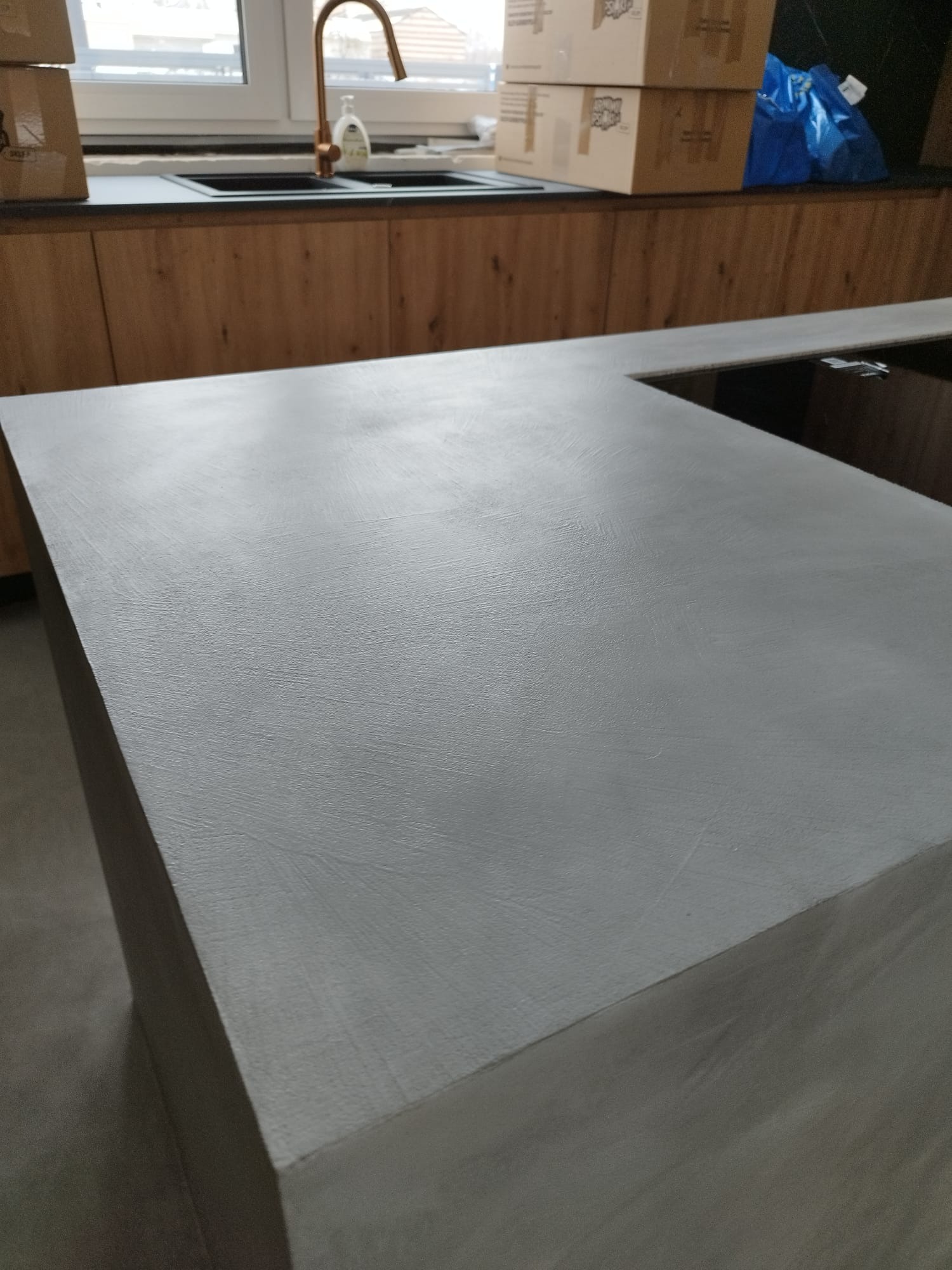 Szara wyspa kuchenna z wykończeniem imitującym beton, widoczna armatura w kolorze miedzi i kartony w tle.