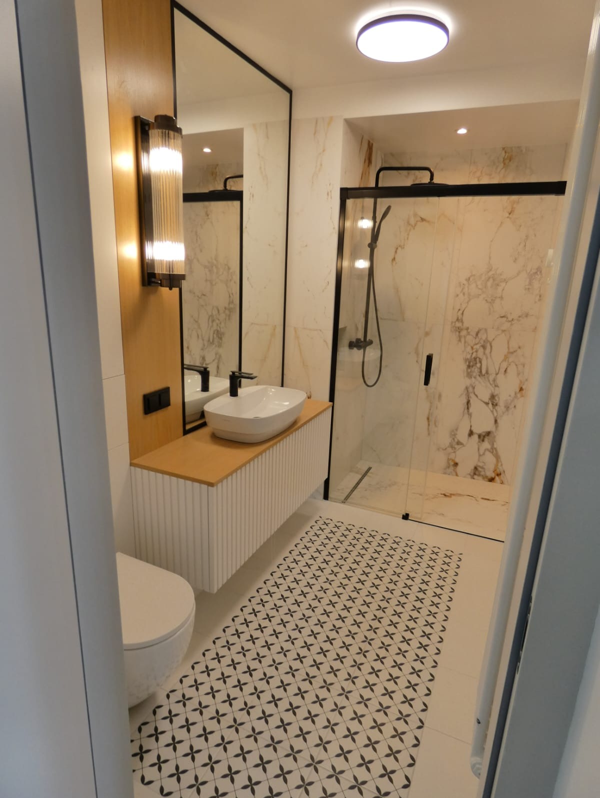 Nowoczesna łazienka z kabiną prysznicową typu walk-in, umywalką nablatową na szafce z ryflowanym frontem, oświetleniem ściennym i podłogą w biało-czarne wzory.