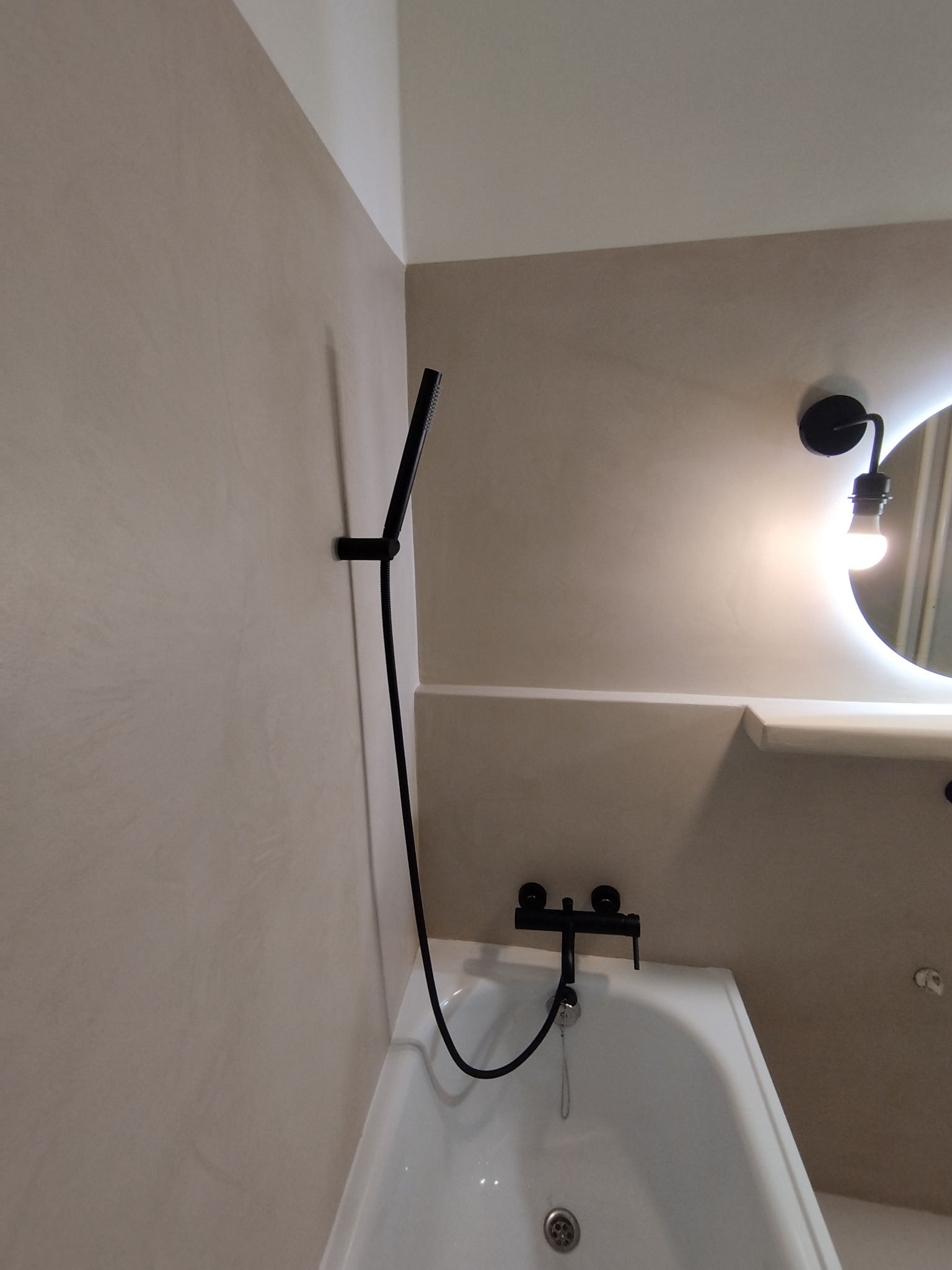 Beżowa łazienka z czarną armaturą: bateria wannowa i deszczownica, ściany wykończone imitacją betonu, okrągłe lustro z podświetleniem LED.