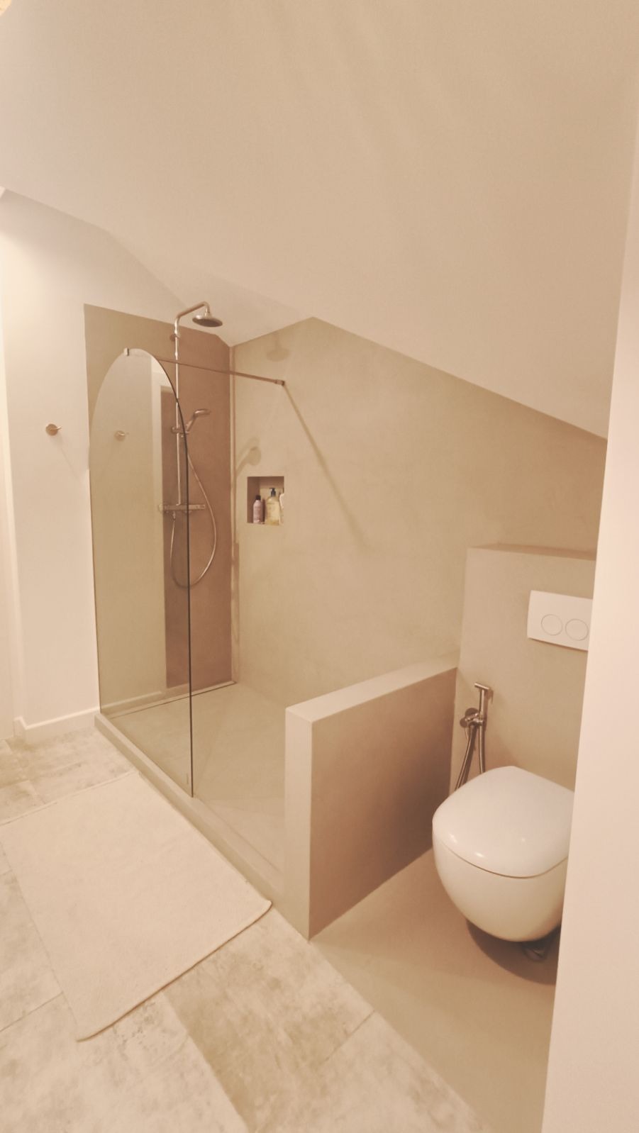 Nowoczesna łazienka na poddaszu z kabiną prysznicową typu walk-in z zaokrąglonym szkłem i toaletą podwieszaną w kolorze beżowym, oświetlona ciepłym światłem.