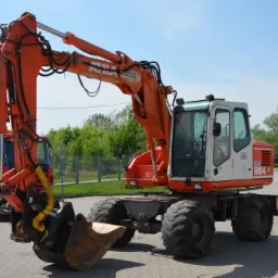 Pomarańczowa koparka kołowa Atlas 1304 na placu budowy, obok czerwona zamiatarka uliczna, w tle zieleń drzew.