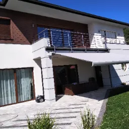 Nowoczesny dom z białą elewacją i brązowymi akcentami, z rozłożoną jasną markizą nad tarasem, widok z poziomu ogrodu na taras z meblami ogrodowymi i fragmentem chodnika z kostki brukowej.