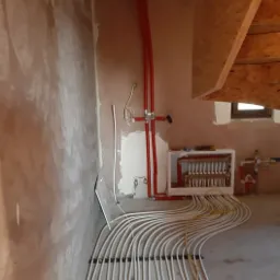 Rozłożone białe rurki instalacji w systemie ogrzewania podłogowego, podłączone do rozdzielacza z czerwonymi rurami w surowym stanie wykończenia pomieszczenia.