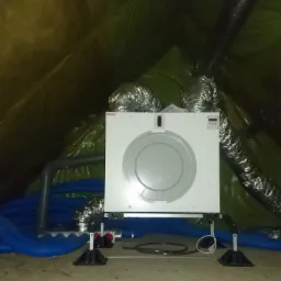 Biała centrala z okrągłym elementem na środku, podłączona do przewodów wentylacyjnych, umieszczona na poddaszu z folią w tle.