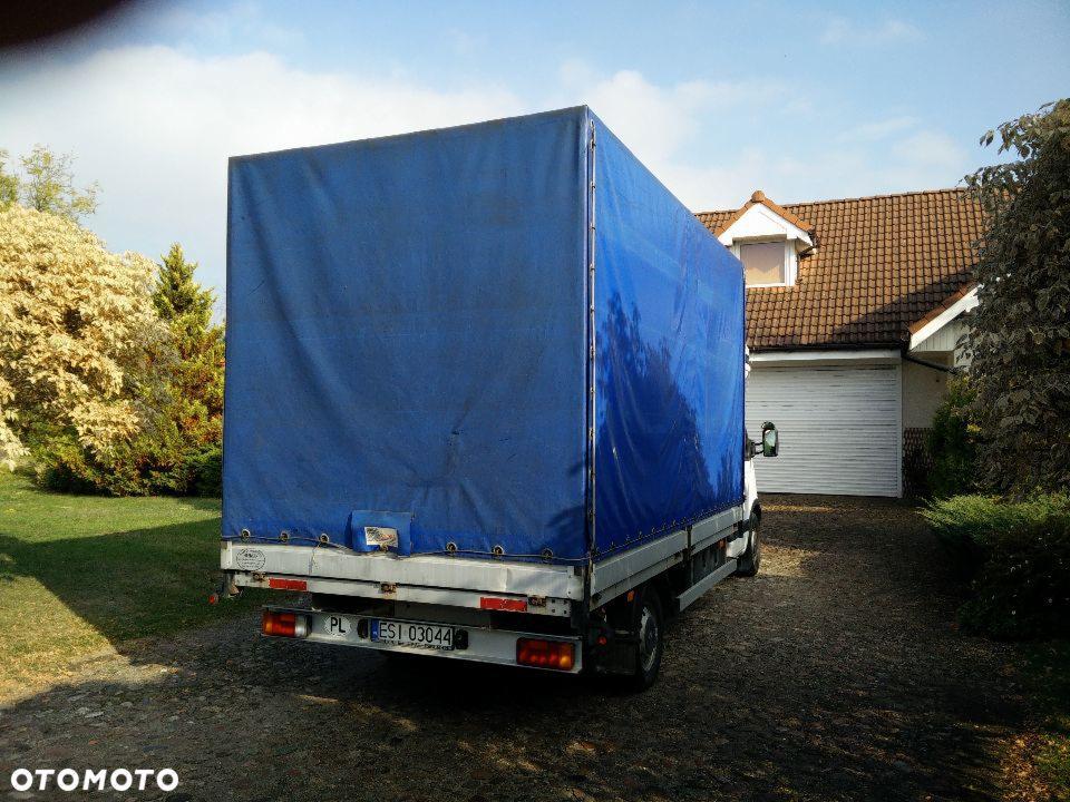 Biały bus z niebieską plandeką zaparkowany na podjeździe z kostki brukowej przed domem z brązową dachówką, widok z tyłu, tablica rejestracyjna PL.