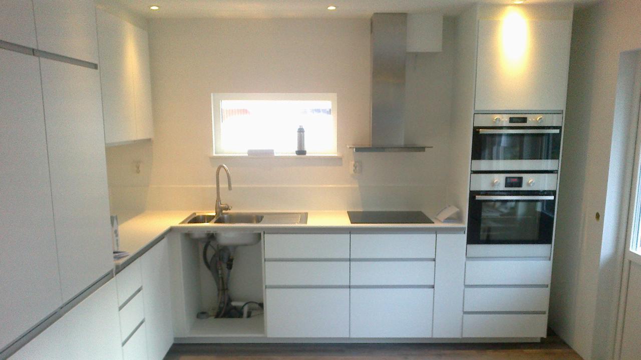 Jasna, minimalistyczna kuchnia z białymi szafkami, stalowym okapem i dwoma piekarnikami w zabudowie. Widok na okno z butelką i gąbką. Umywalka z widocznymi rurami.