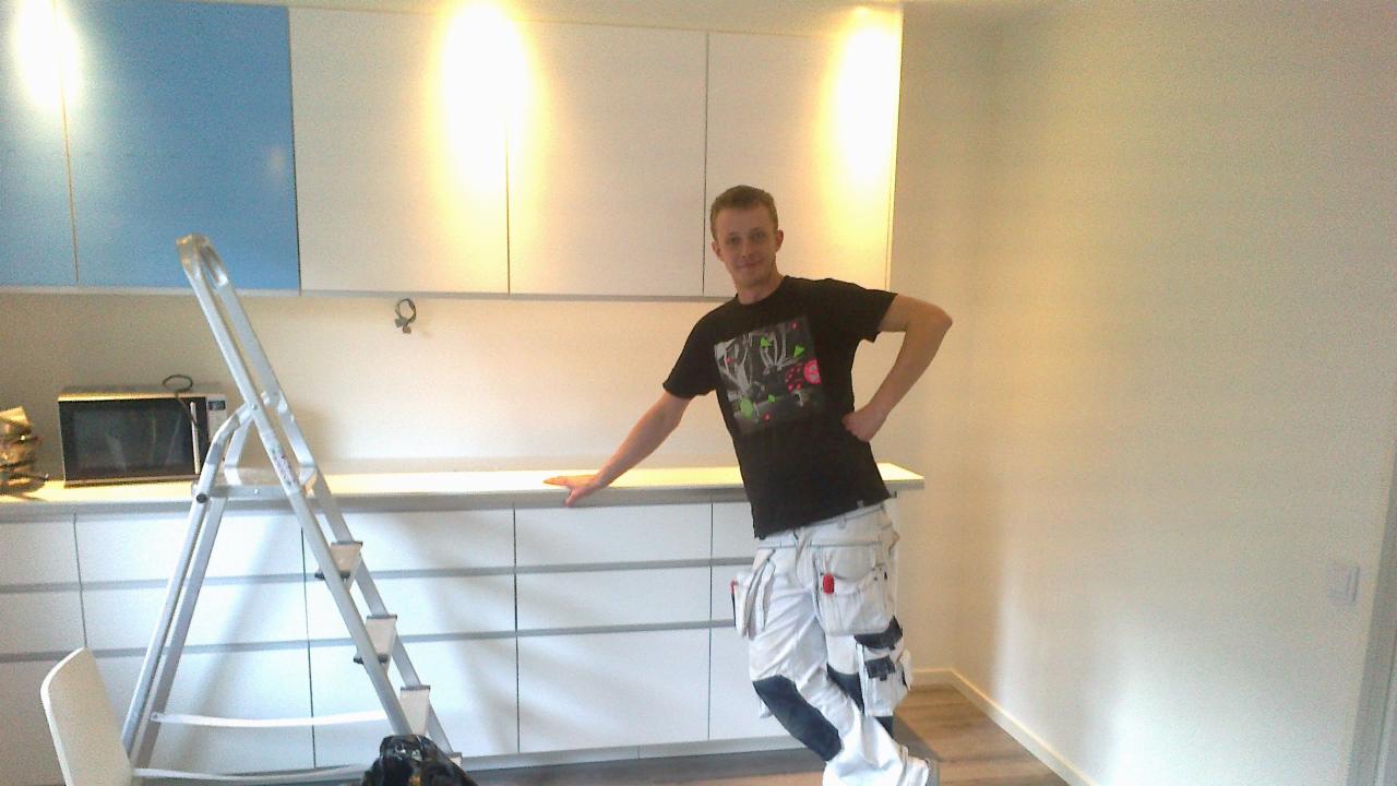 Remont kuchni w Szymbarku: mężczyzna w spodniach roboczych stoi obok drabiny na tle szafek kuchennych z niebieskim akcentem. Świeżo pomalowane ściany.