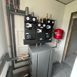 Zmontowana instalacja grzewcza z szarą obudową kotła AFRISO, izolowanymi rurami, zaworami i manometrami w pomieszczeniu z szarymi płytkami podłogowymi i fragmentem surowej ściany.