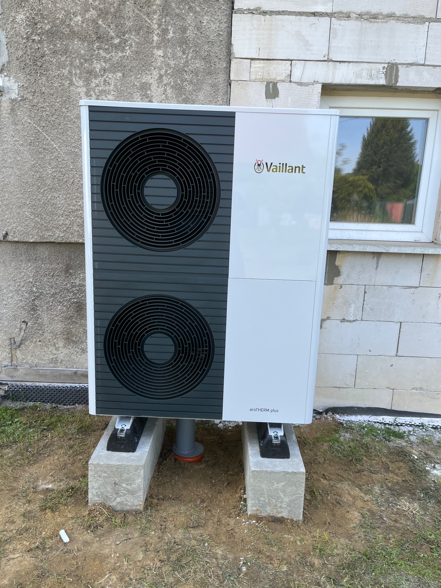 Biała pompa ciepła marki Vaillant, model aroTHERM plus, zamontowana na betonowych podstawach przed budynkiem z pustaków, widok z przodu, okno odbija drzewa.