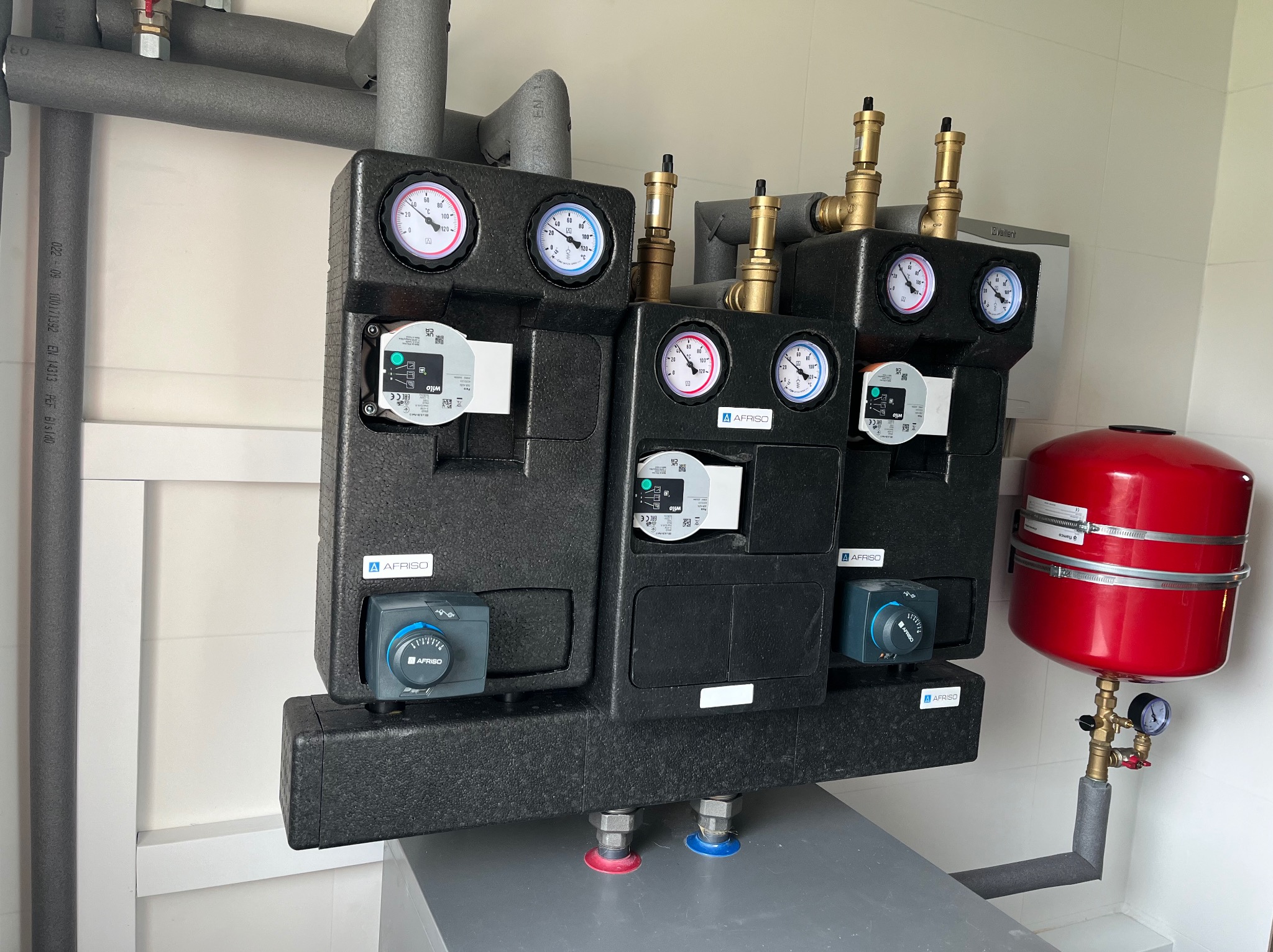 Dwa moduły mieszające z izolacją termiczną, manometrami i pompami obiegowymi Wilo, zamontowane na szarej podstawie w pomieszczeniu z białymi płytkami, widoczny czerwony zbiornik wyrównawczy, rury...