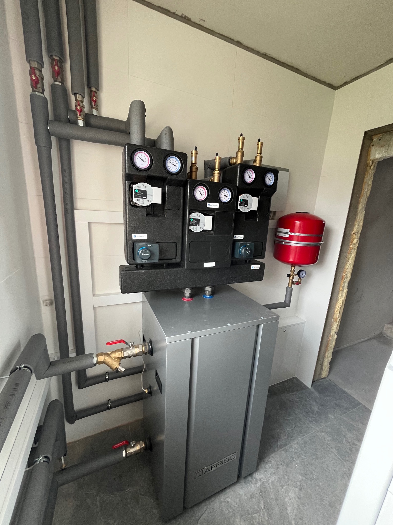 Zmontowana instalacja grzewcza z szarą obudową kotła AFRISO, izolowanymi rurami, zaworami i manometrami w pomieszczeniu z szarymi płytkami podłogowymi i fragmentem surowej ściany.