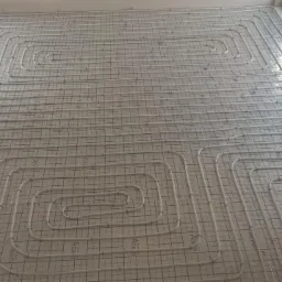 Instalacja ogrzewania podłogowego w jasnym pomieszczeniu z oknami. Białe rury ułożone na siatce, widoczne ściany w stanie surowym. Altech.