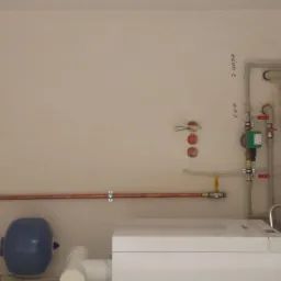 Nowa instalacja CO/CWU z miedzianymi rurami, zbiornikiem i piecem. Widoczne elementy przyłączeniowe, zawory i manometr. Ściany w jasnym kolorze, brak dekoracji.