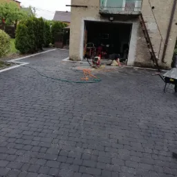 Ułożona, grafitowa kostka brukowa przed budynkiem z garażem. Brukarz w trakcie pracy, widoczna zagęszczarka i narzędzia. Obramowanie z białej kostki.