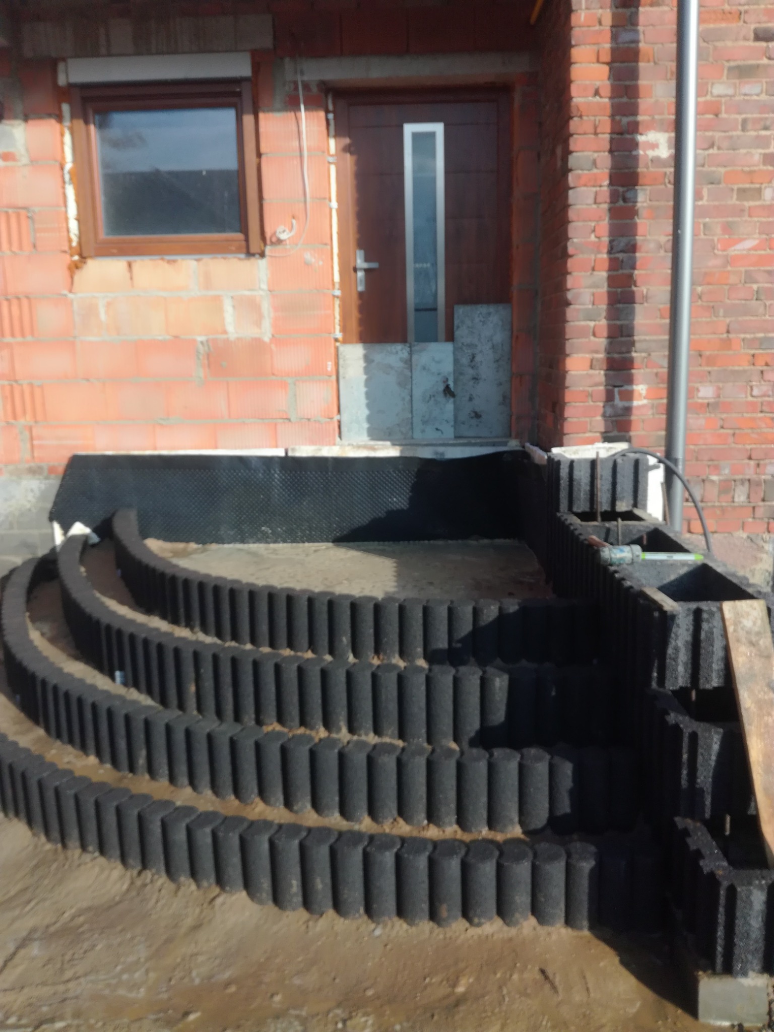 Montaż schodów z obrzeży trawnikowych przed wejściem do budynku z cegły. Widoczne narzędzia i materiały przygotowane do dalszych prac.