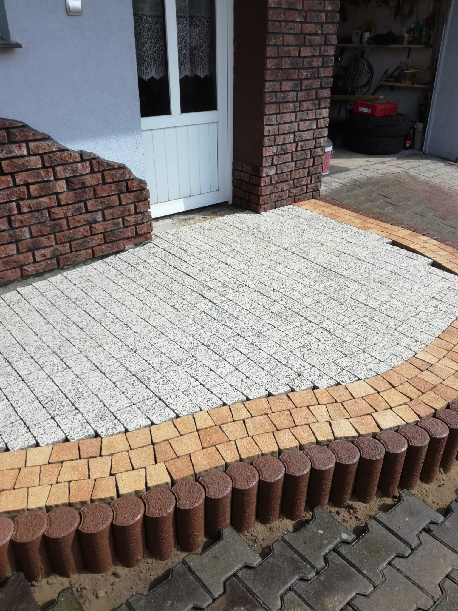 Wykończone wejście do domu w Gostyniu z kostki brukowej w odcieniach szarości i brązu, ułożonej w dekoracyjny wzór. Widoczne obrzeża i fragment podjazdu.