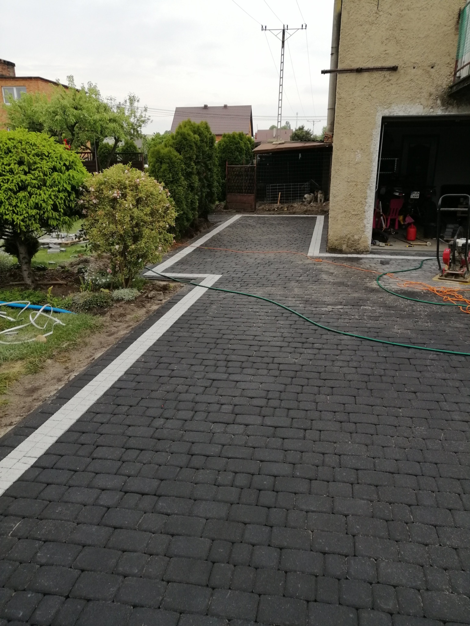 Ułożona, czarna kostka brukowa z białymi obrzeżami na podjeździe w Gostyniu. Widoczny ogród i budynek gospodarczy.