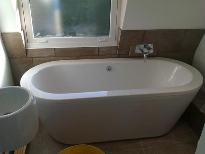 Biała, owalna wanna wolnostojąca z chromowaną baterią, obudowana brązowymi płytkami, ustawiona pod oknem w jasnej łazience. Umywalka obok wanny.