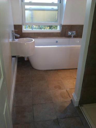 Jasna łazienka po remoncie: biała wanna i umywalka na tle okna, brązowe płytki na podłodze i ścianie. Widok zza otwartych drzwi.