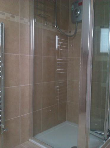 Kabina prysznicowa z beżowymi płytkami, widoczny grzejnik drabinkowy i podgrzewacz wody. Ujęcie z bliska, prezentujące detale wykończenia łazienki.