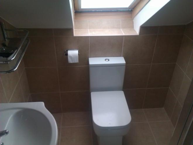 Beżowa, mała łazienka na poddaszu z toaletą, umywalką i grzejnikiem. Ściany i podłoga wyłożone kwadratowymi płytkami. Okno dachowe w tle.