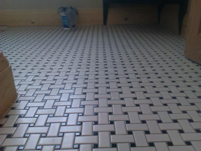 Beżowa podłoga z płytek ułożonych w wzór koszykowy z czarnymi akcentami, widok z dołu, fragment szafki w tle i listwa przypodłogowa.