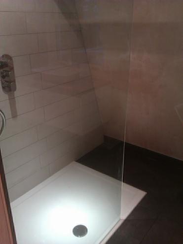 Nowoczesna kabina prysznicowa z białym brodzikiem i szklaną ścianką, wykończona jasnymi, prostokątnymi płytkami na ścianie i ciemną podłogą.