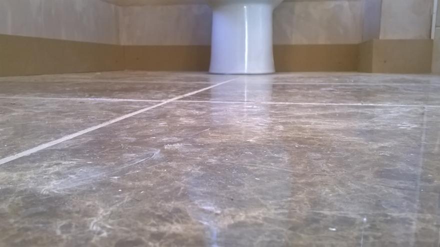 Szare płytki podłogowe z białymi fugami w łazience, widoczna dolna część toalety i fragment ściany z brązowym cokołem. Płytki z delikatnym wzorem.