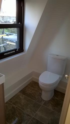 Biała toaleta w małej łazience na poddaszu z brązowymi płytkami podłogowymi. Okno z ciemną ramą. Świeżo pomalowane ściany na biało.